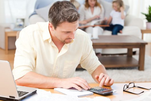 Man calculating his bills while his family are sitting on the sofa